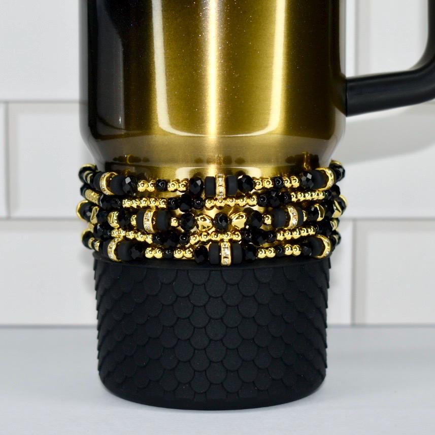 A gold-colored travel mug with a black handle. The mug is adorned with multiple layers of black and gold beaded accents. The bottom of the mug features a textured black rubber sleeve.