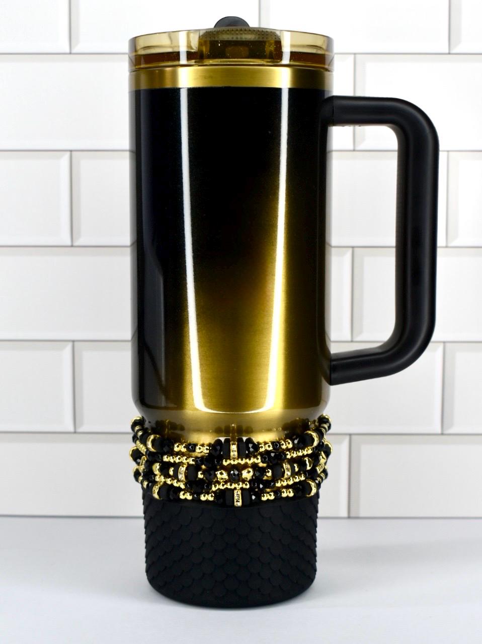 A black and gold Stanley with a black handle. The mug has a gold lid and a decorative band of black and gold beads. The bottom of the mug has a textured black base.