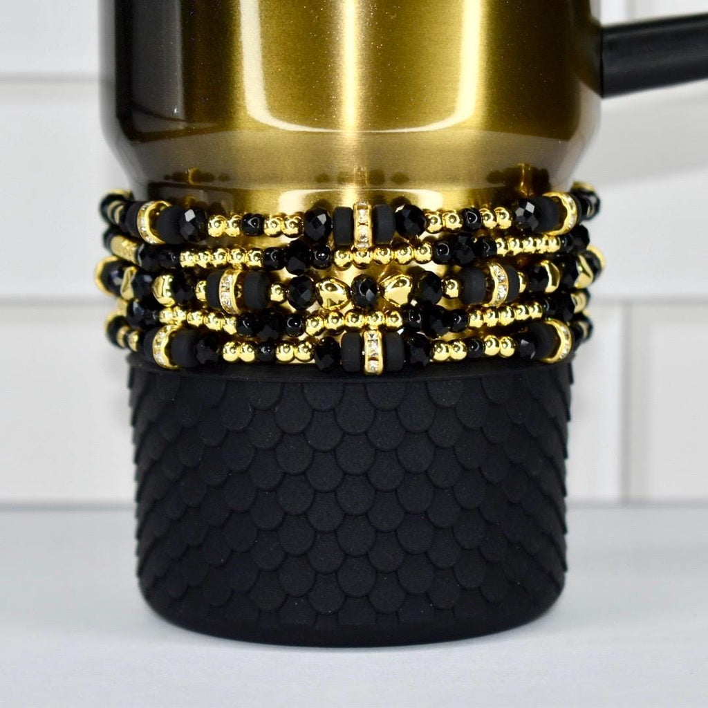 A Black Honey Stanley tumbler with a black textured base and several beaded bracelets. The bracelets feature black beads with gold accents and small, clear embellishments. The tumbler has a black handle.