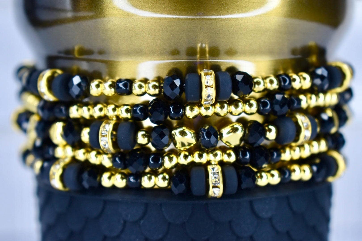 Stack of beaded bracelets in black and gold. The bracelets feature a mix of black faceted beads, small gold beads, and gold-toned spacers with clear rhinestones. The bracelets are stacked on a black textured surface.