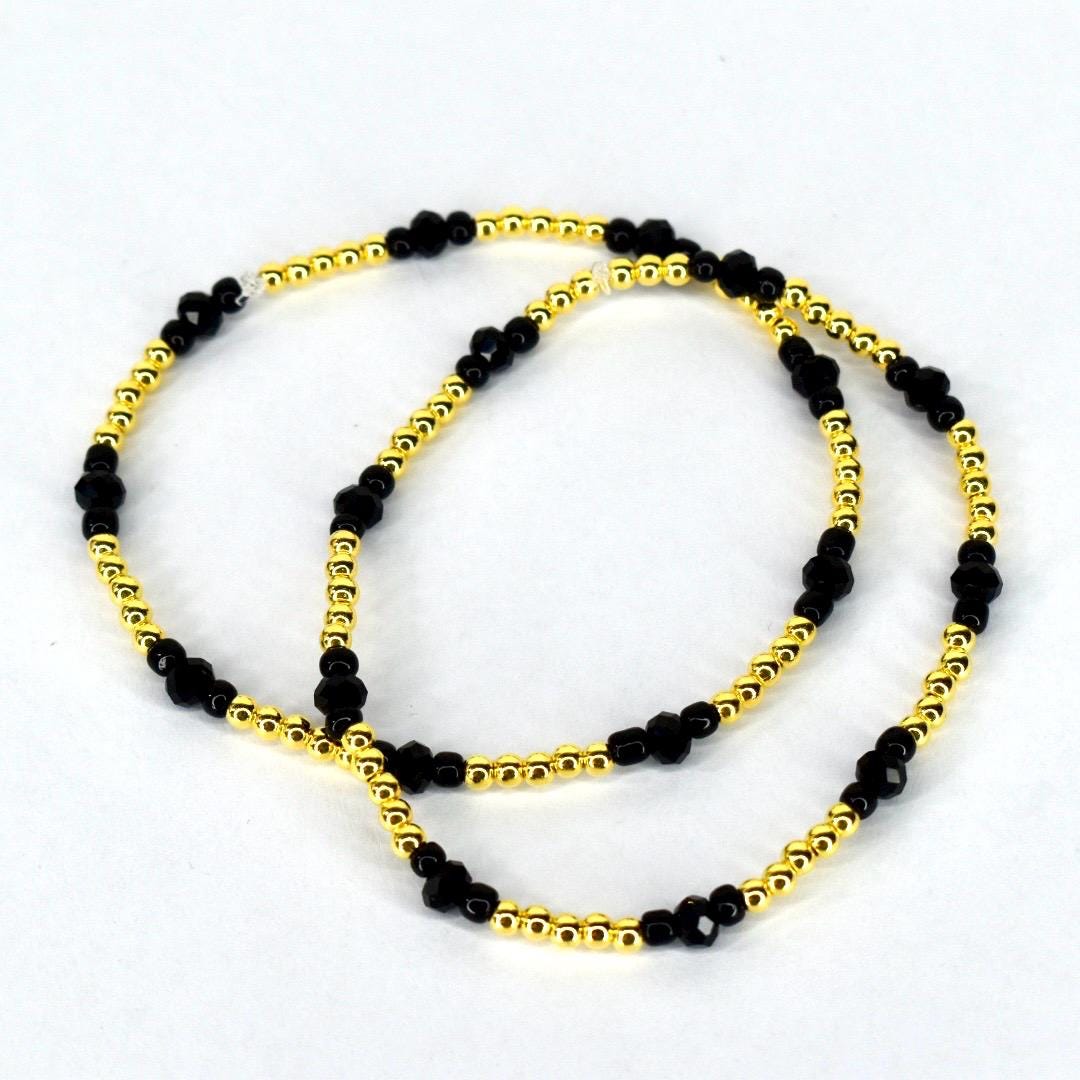 Two beaded bracelets. The bracelets feature a combination of small, gold-colored beads and larger, faceted black beads. The bracelets are displayed on a white background.