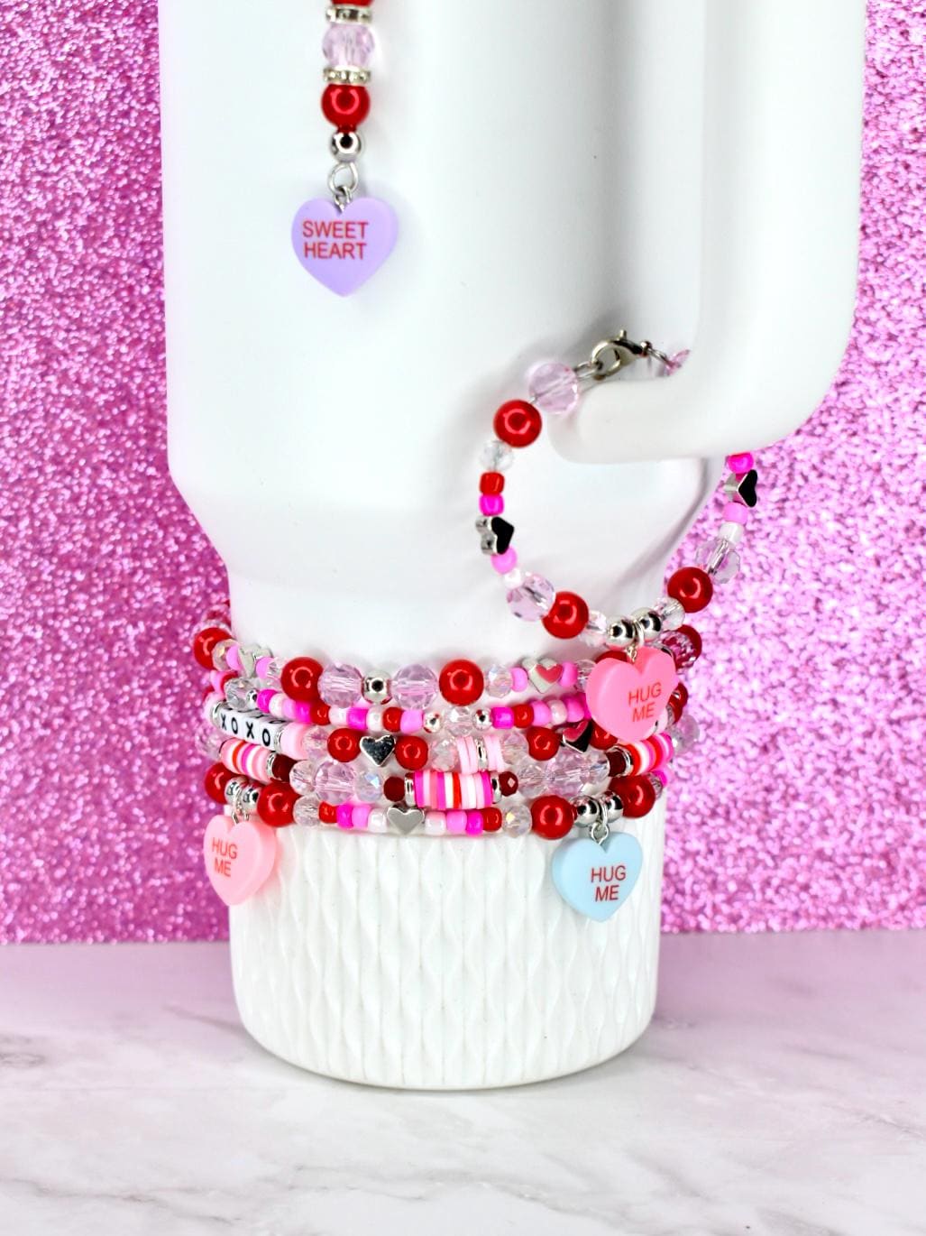 Stack of beaded bracelets with heart charms and the words "HUG ME" and "XOXO". The bracelets are red, pink, and clear with silver accents. A matching beaded necklace with a "SWEET HEART" charm is also visible.