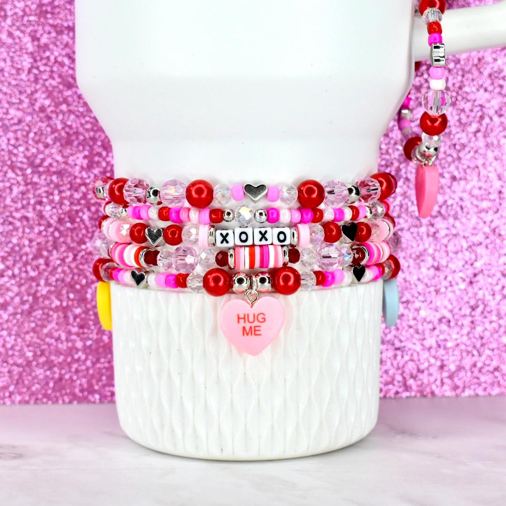 A stack of beaded bracelets in shades of red, pink, and clear, with heart and XOXO accents. One bracelet has a pink heart charm that says "HUG ME". The bracelets are stacked on a white mug.