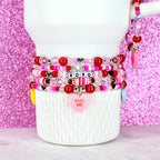 A stack of beaded bracelets in shades of red, pink, and clear, with heart and XOXO accents. One bracelet has a pink heart charm that says "HUG ME". The bracelets are stacked on a white mug.