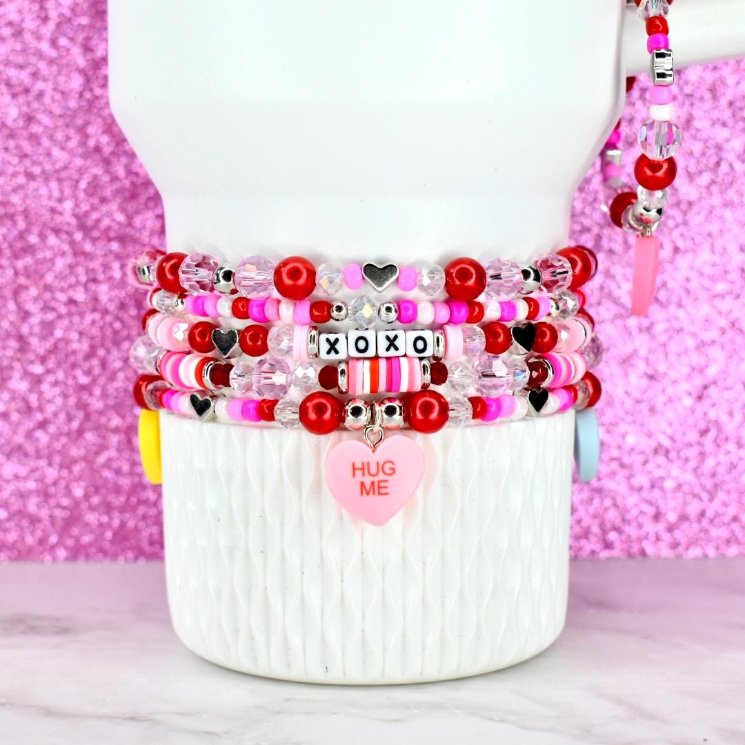 Stack of beaded bracelets in shades of pink, red, and white. The bracelets feature heart-shaped beads, clear beads, and beads spelling "XOXO". A pink heart charm with "HUG ME" is attached to one bracelet.