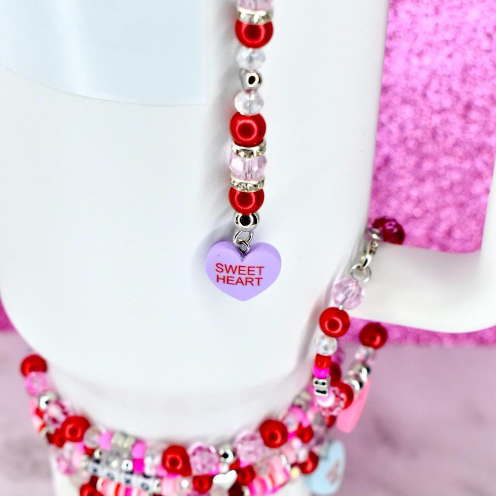 A white mug with a beaded charm. The charm features red, pink, and clear beads, with a purple heart pendant that reads "Sweet Heart". The mug is set against a pink background.