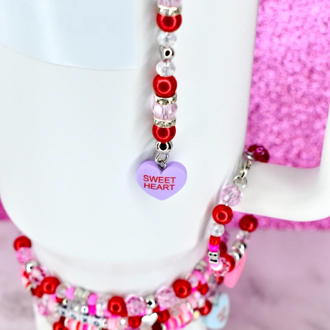 A white mug with a beaded charm. The charm features red, pink, and clear beads, with a purple heart pendant that reads "Sweet Heart". The mug is set against a pink background.
