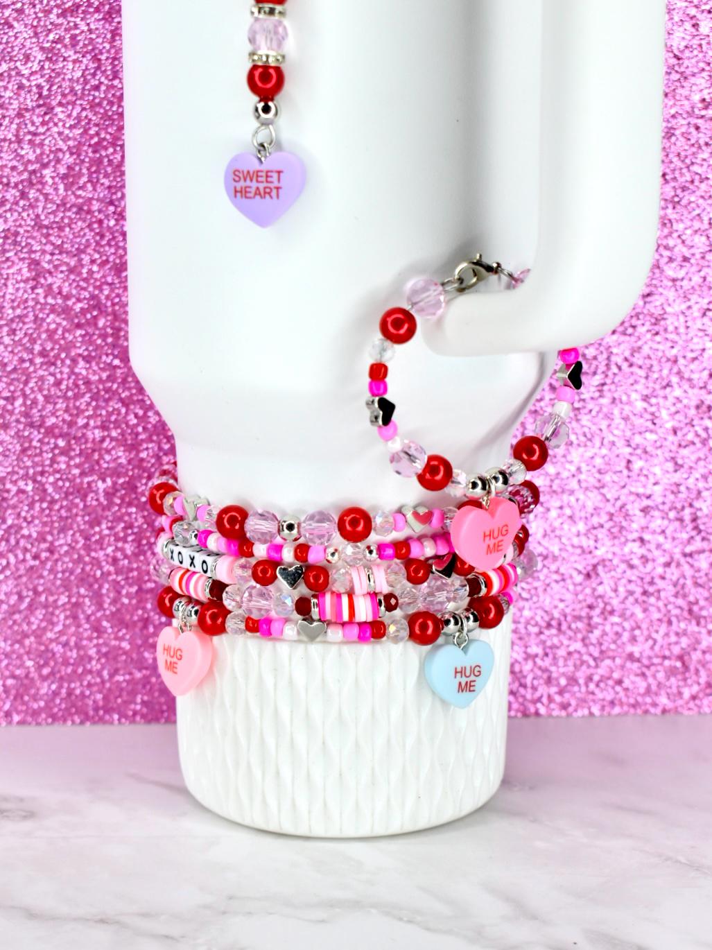 A collection of beaded bracelets and a matching earring set. The bracelets feature red, pink, and clear beads, with heart-shaped charms that read "HUG ME" and "XOXO". The earring has a purple heart charm that reads "SWEET HEART".