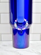 A blue tumbler with a beaded bracelet. The bracelet features blue, clear, and pink beads, and a small star charm. The tumbler has a gradient finish, transitioning from blue to purple.