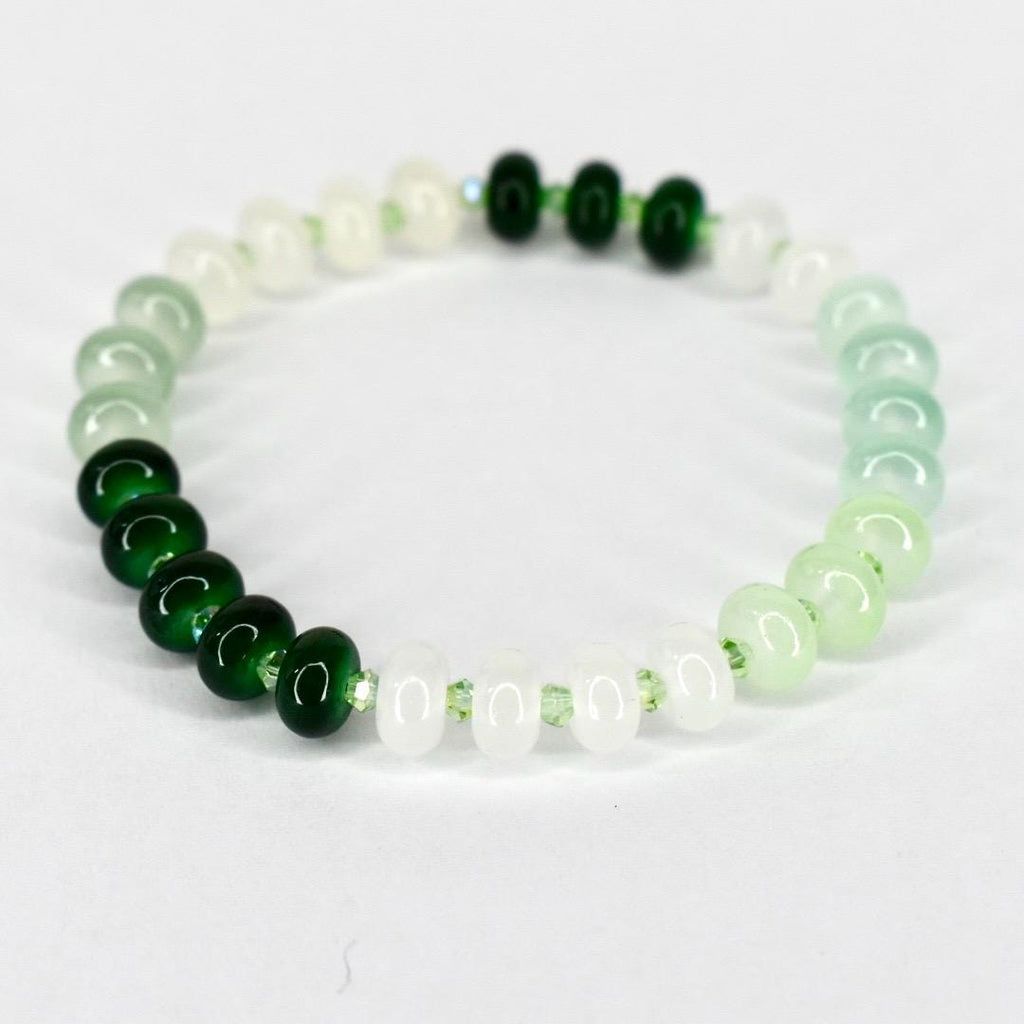 A beaded bracelet featuring a gradient of green and white beads. The bracelet transitions from dark green to light green and white, with a few dark green beads clustered together. The beads are round and strung on a clear elastic cord.