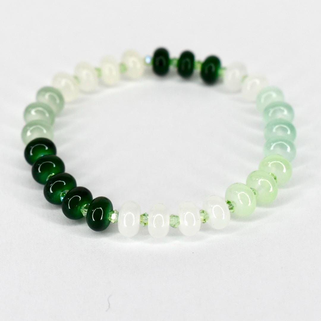 A beaded bracelet featuring a gradient of green and white beads. The bracelet transitions from dark green to light green and white, with a few dark green beads clustered together. The beads are round and strung on a clear elastic cord.
