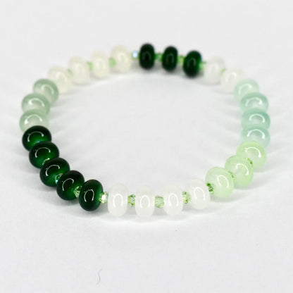 A beaded bracelet featuring a gradient of green and white beads. The bracelet transitions from dark green to light green and white, with a few dark green beads clustered together. The beads are round and strung on a clear elastic cord.