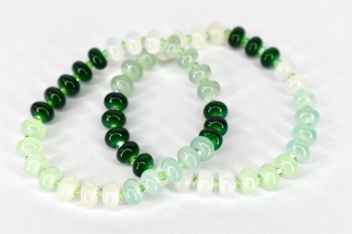 Two beaded bracelets with a gradient of colors. The beads transition from white to light green to dark green. The bracelets are strung on a thin, light green cord. The beads are oval-shaped.
