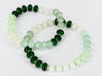 Two beaded bracelets with a gradient of green and white beads. The beads transition from dark green to light green and then to white. The bracelets are strung on a clear, flexible cord.