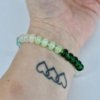 A beaded bracelet with a mix of light green, white, and dark green beads. The bracelet is strung on a light green cord and is worn on a wrist. The beads are round and oval-shaped.