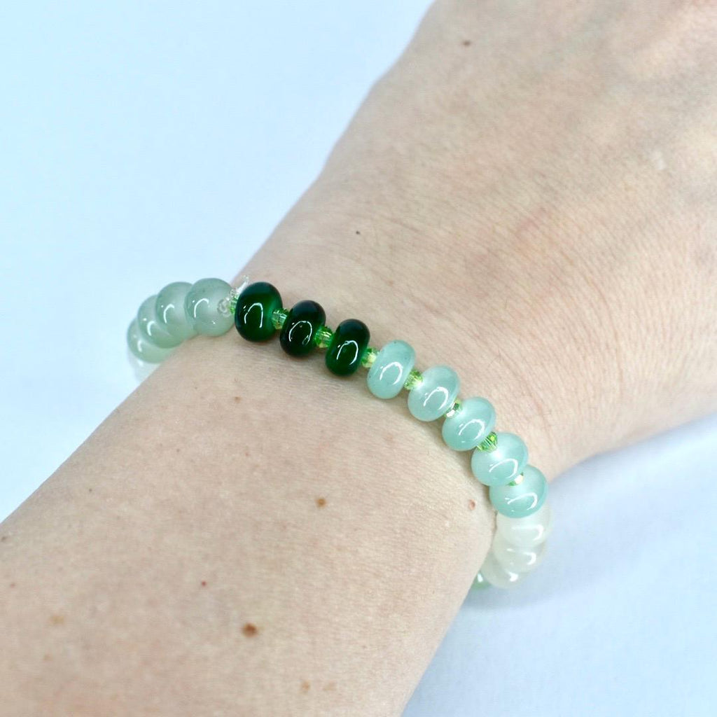 A beaded bracelet featuring a mix of green and white beads. The bracelet has a gradient effect, transitioning from white to light green and then to darker green beads. The beads are strung on a clear elastic cord.