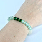 A beaded bracelet featuring a mix of green and white beads. The bracelet has a gradient effect, transitioning from white to light green and then to darker green beads. The beads are strung on a clear elastic cord.