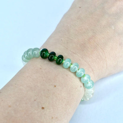 A beaded bracelet featuring a mix of green and white beads. The bracelet has a gradient effect, transitioning from white to light green and then to darker green beads. The beads are strung on a clear elastic cord.