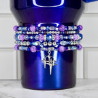 A deep blue tumbler adorned with multiple beaded bracelets in shades of purple, blue, and silver. The bracelets feature various bead shapes and sizes, with a silver charm of a starburst design hanging from the bottom.