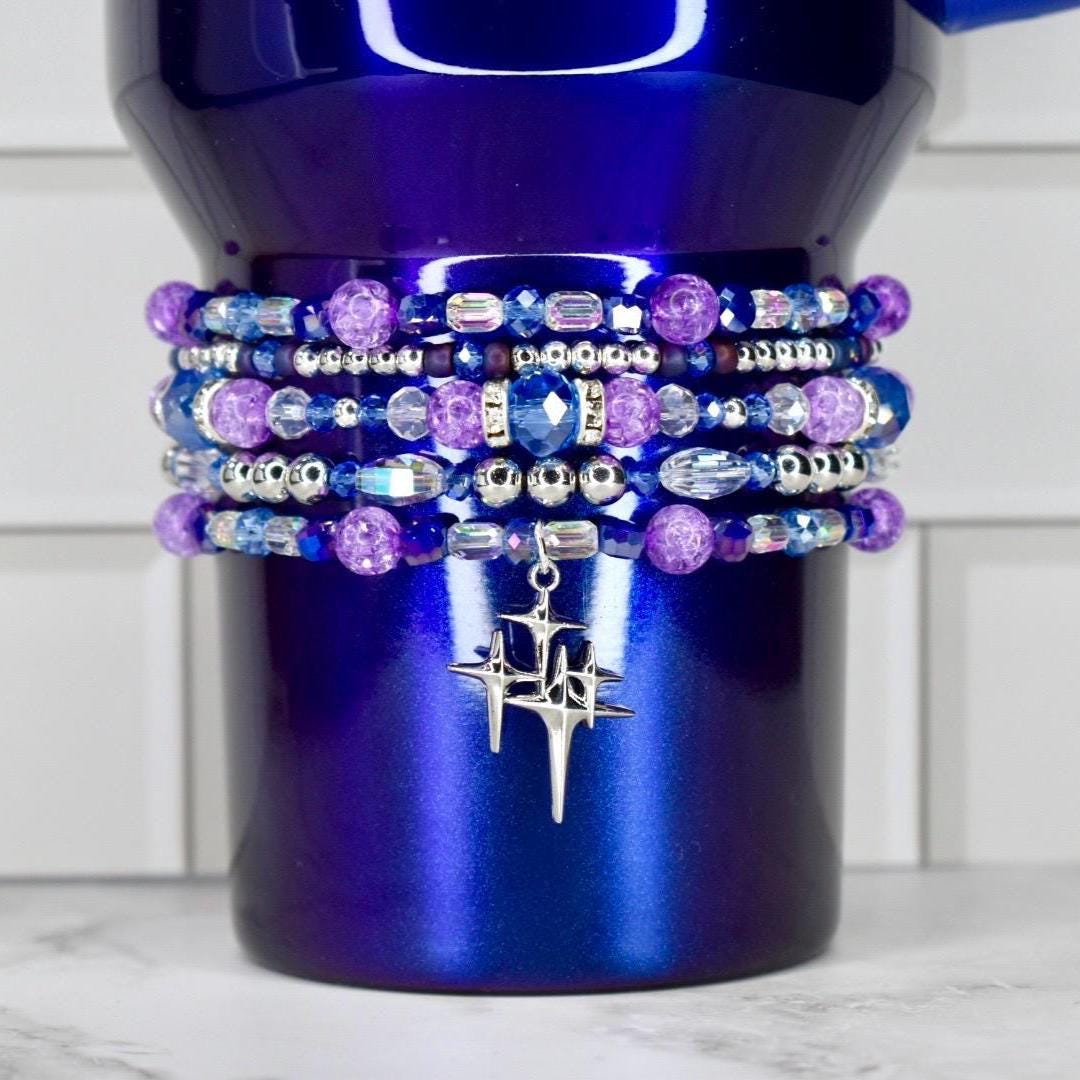 A deep blue tumbler adorned with multiple beaded bracelets in shades of purple, blue, and silver. The bracelets feature various bead shapes and sizes, with a silver charm of a starburst design hanging from the bottom.