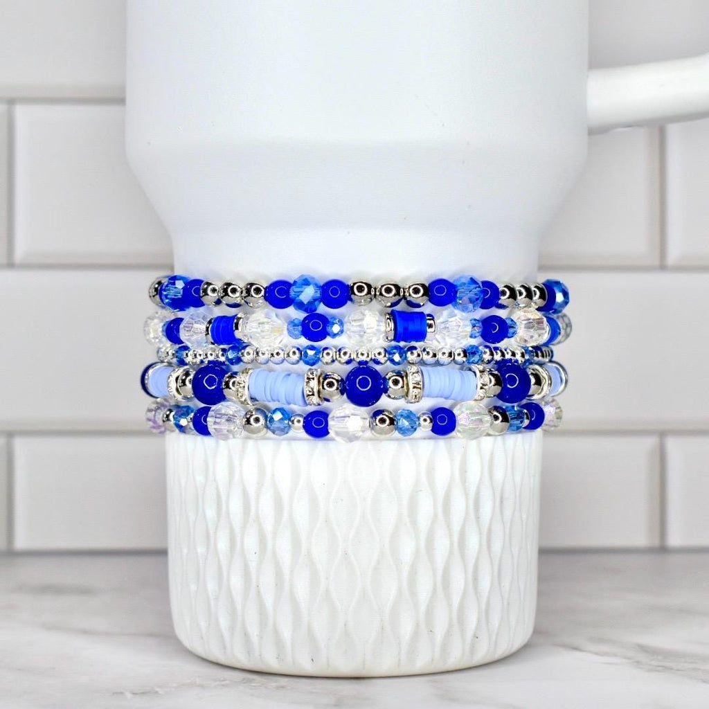 A stack of five beaded bracelets in shades of blue and clear, with silver accents. The bracelets are displayed on a white, textured cup. The beads vary in size and shape, creating a layered effect.