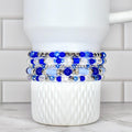 A stack of five beaded bracelets in shades of blue and clear, with silver accents. The bracelets are displayed on a white, textured cup. The beads vary in size and shape, creating a layered effect.