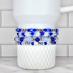 A stack of five beaded bracelets in shades of blue and clear, with silver accents. The bracelets are displayed on a white, textured cup. The beads vary in size and shape, creating a layered effect.
