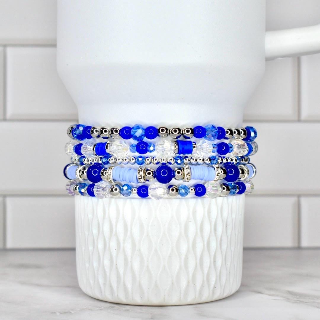 A stack of five beaded bracelets in shades of blue and clear, with silver accents. The bracelets are displayed on a white, textured cup. The beads vary in size and shape, creating a layered effect.