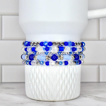 A stack of five beaded bracelets in shades of blue and clear, with silver accents. The bracelets are displayed on a white, textured cup. The beads vary in size and shape, creating a layered effect.