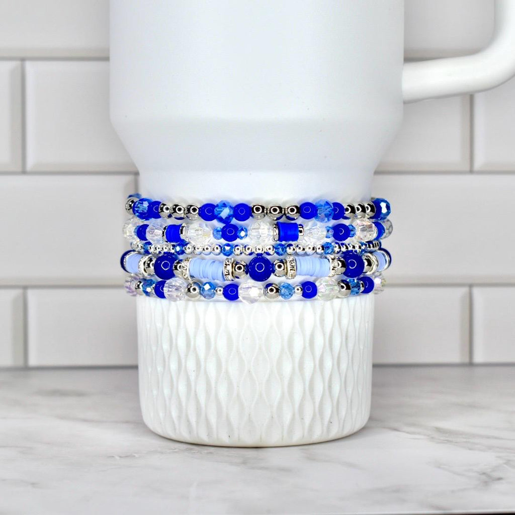 A stack of five beaded bracelets in shades of blue and clear, with silver accents. The bracelets are displayed on a white, textured ceramic mug. The beads vary in size and shape, creating a layered effect.