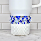 A stack of five beaded bracelets in shades of blue and clear, with silver accents. The bracelets are displayed on a white, textured ceramic mug. The beads vary in size and shape, creating a layered effect.