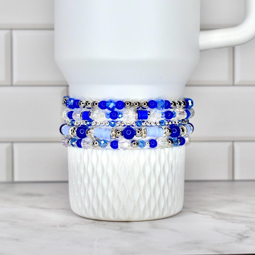 A stack of five beaded bracelets in shades of blue and clear, with silver accents. The bracelets are displayed on a white, textured ceramic mug. The beads vary in size and shape, creating a layered effect.