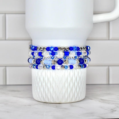A stack of five beaded bracelets in shades of blue and clear, with silver accents. The bracelets are displayed on a white, textured ceramic mug. The beads vary in size and shape, creating a layered effect.