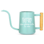 A light teal watering can with a long, curved spout and a wooden handle. The can features a white floral design and the text "Love grows here Est. 2025".