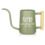 Sage green watering can with a long spout and wooden handle. The can features a white floral design and the text "love grows here Est. 2025". A gardening tool for indoor or outdoor use.