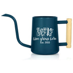 A dark blue watering can with a long, curved spout and a wooden handle. The can features a white floral design and the text "love grows here Est. 2025".