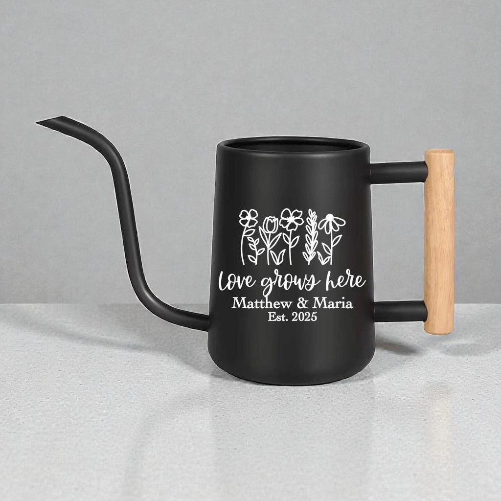 A black watering can with a long, curved spout and a wooden handle. The can features white text that reads "Love grows here" with the names "Matthew & Maria" and the year "Est. 2025" below a floral design.