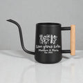 A black watering can with a long, curved spout and a wooden handle. The can features white text that reads "Love grows here" with the names "Matthew & Maria" and the year "Est. 2025" below a floral design.