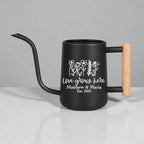 A black watering can with a long, curved spout and a wooden handle. The can features white text that reads "Love grows here" with the names "Matthew & Maria" and the year "Est. 2025" below a floral design.