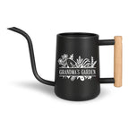 Black metal watering can with a long, curved spout and a wooden handle. The can features the text "Grandma's Garden" in white, along with floral and gardening tool illustrations. The handle is made of light-colored wood.