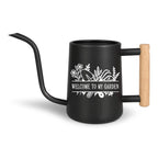 A black metal watering can with a long, curved spout and a wooden handle. The can features a white floral design and the text "WELCOME TO MY GARDEN". Ideal for gardening and watering plants.