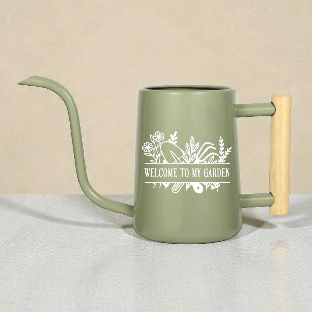 Sage green watering can with a long spout and wooden handle. The can features a white floral design and the text "WELCOME TO MY GARDEN". The watering can is designed for indoor or outdoor use.