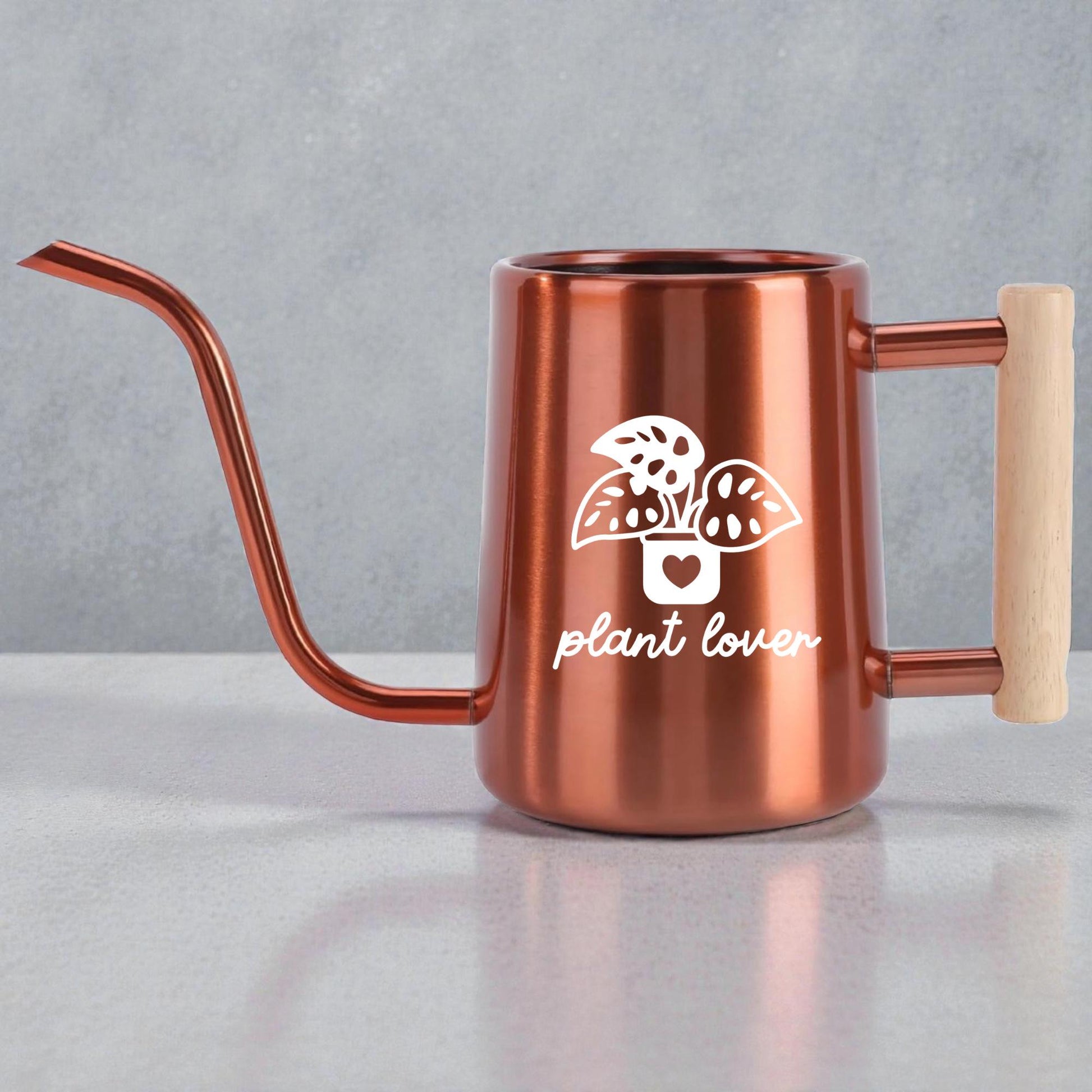 Copper watering can with a long, slender spout and a wooden handle. The can features a white graphic of a potted plant with the words "plant lover" below it. The watering can is designed for indoor or outdoor plant care.