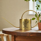 A gold-colored watering can with a long spout and a circular handle. The can features a black floral design and the name "Amanda" in script. It sits on a wooden table, suggesting a gardening theme.
