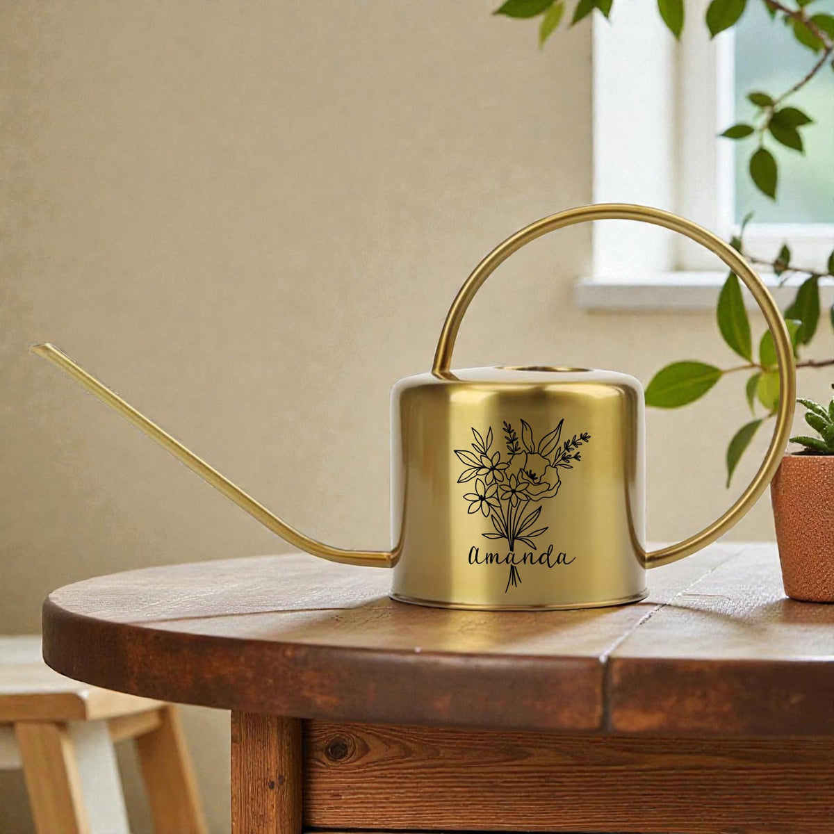 A gold-colored watering can with a long spout and a circular handle. The can features a black floral design and the name "Amanda" in script. It sits on a wooden table, suggesting a gardening theme.
