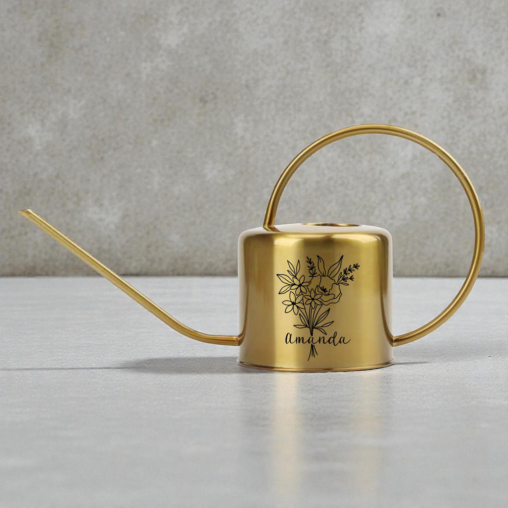 A gold-colored metal watering can with a long spout and a circular handle. The can features a black floral design and the name "Amanda" in script. The watering can is designed for indoor or outdoor plant care.