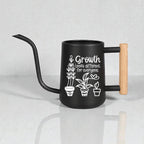 Black gooseneck watering can with wood handle. Three house plants are featured as part of the design on the side of the watering can along with the words 'growth looks different for everyone.'