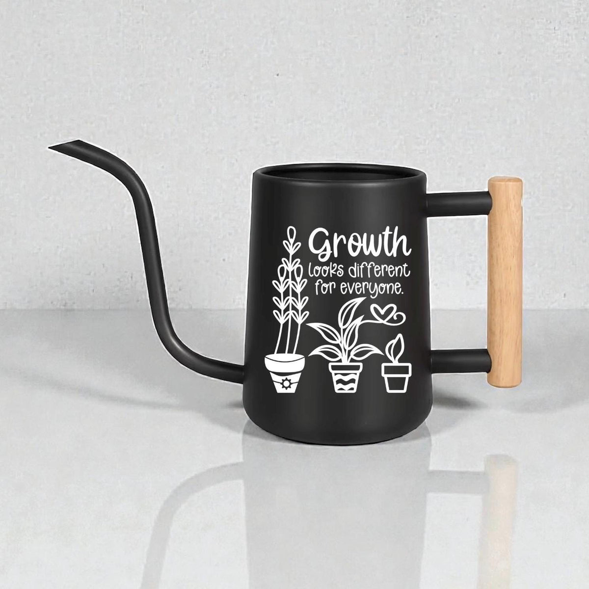 Black gooseneck watering can with wood handle. Three house plants are featured as part of the design on the side of the watering can along with the words 'growth looks different for everyone.'