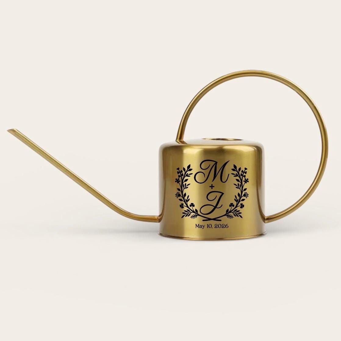 A gold-colored metal watering can with a long spout and a circular handle. The can features a black floral wreath design with the initials "M + J" and the date "May 10, 2026" printed below the initials.
