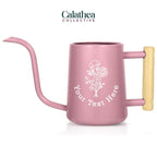 A light pink watering can with long gooseneck spout and wood handle with a white flower bouquet design and 'your text here' below the bouquet in white permanent vinyl.